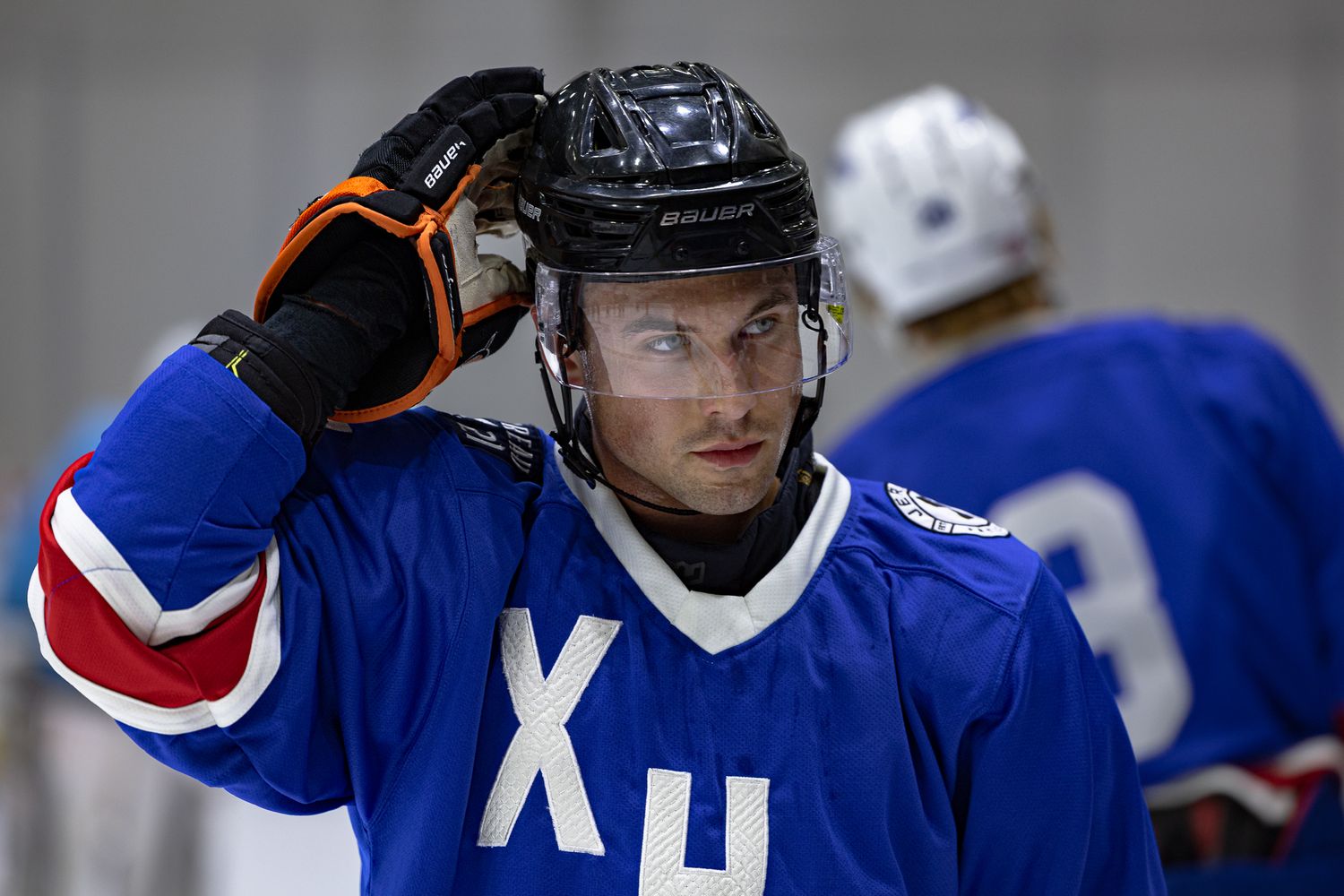 Hockey player in blue jersey with Cyrillic letters adjusts helmet on ice rink during practice.