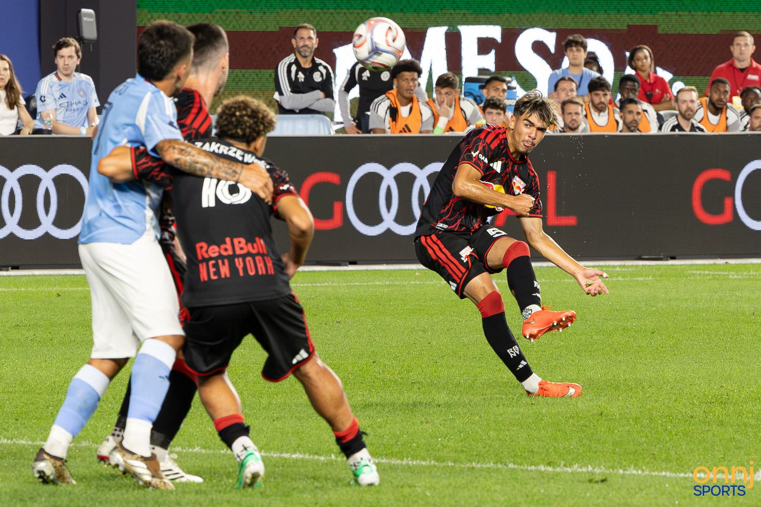 Red Bulls and NYCFC players compete in an intense MLS soccer match, with a player striking the ball mid-air.