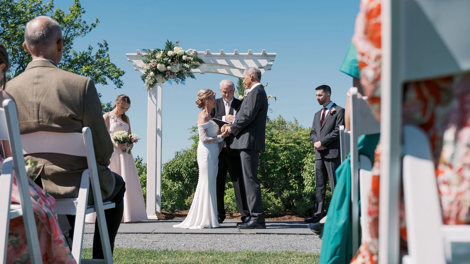 Wide over-the-shoulder perspective of a wedding ceremony at The Boathouse, capturing the wedding party and guests under a clear blue New Eng