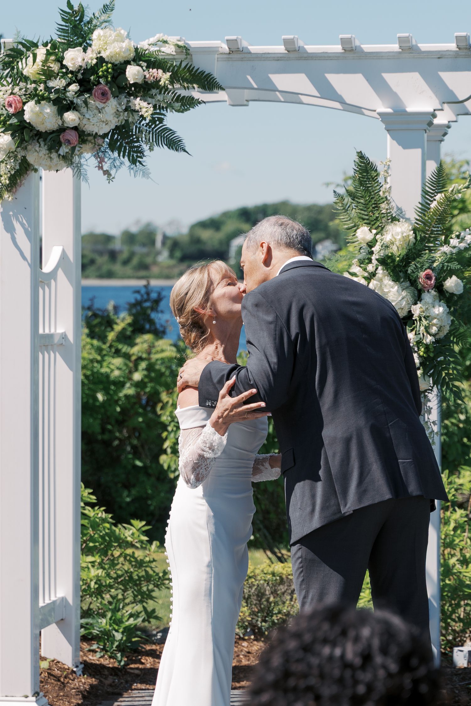 Bride and groom’s first kiss under a white floral-adorned pergola during a waterfront ceremony in Tiverton, RI.