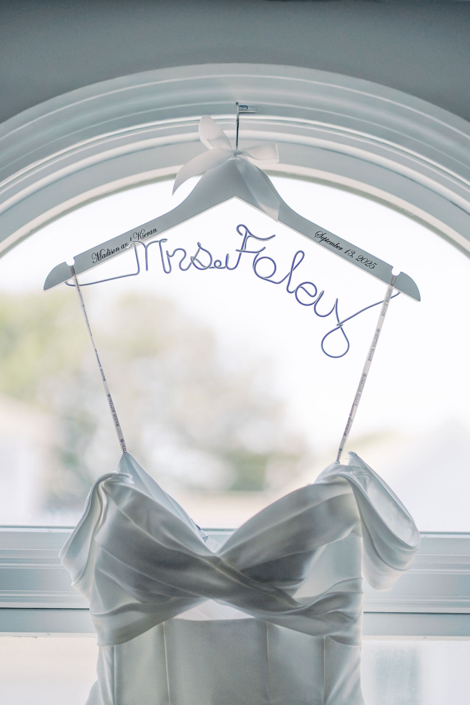 Custom 'Mrs. Foley' wire wedding hanger holding a classic satin bridal gown in front of an arched window.