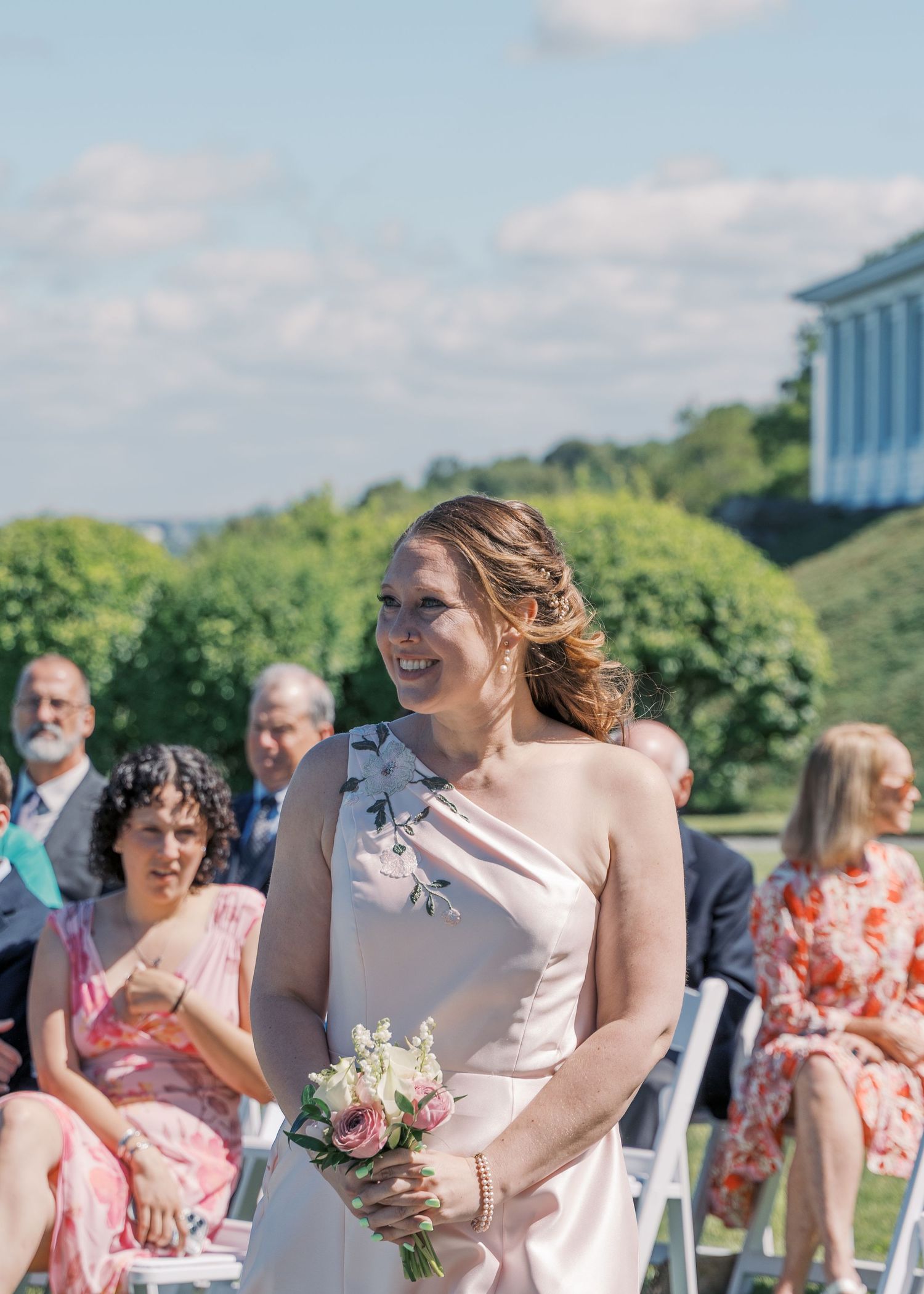 A smiling bridesmaid in a blush floral dress looking on during a sunny outdoor wedding ceremony at The Boathouse Tiverton.