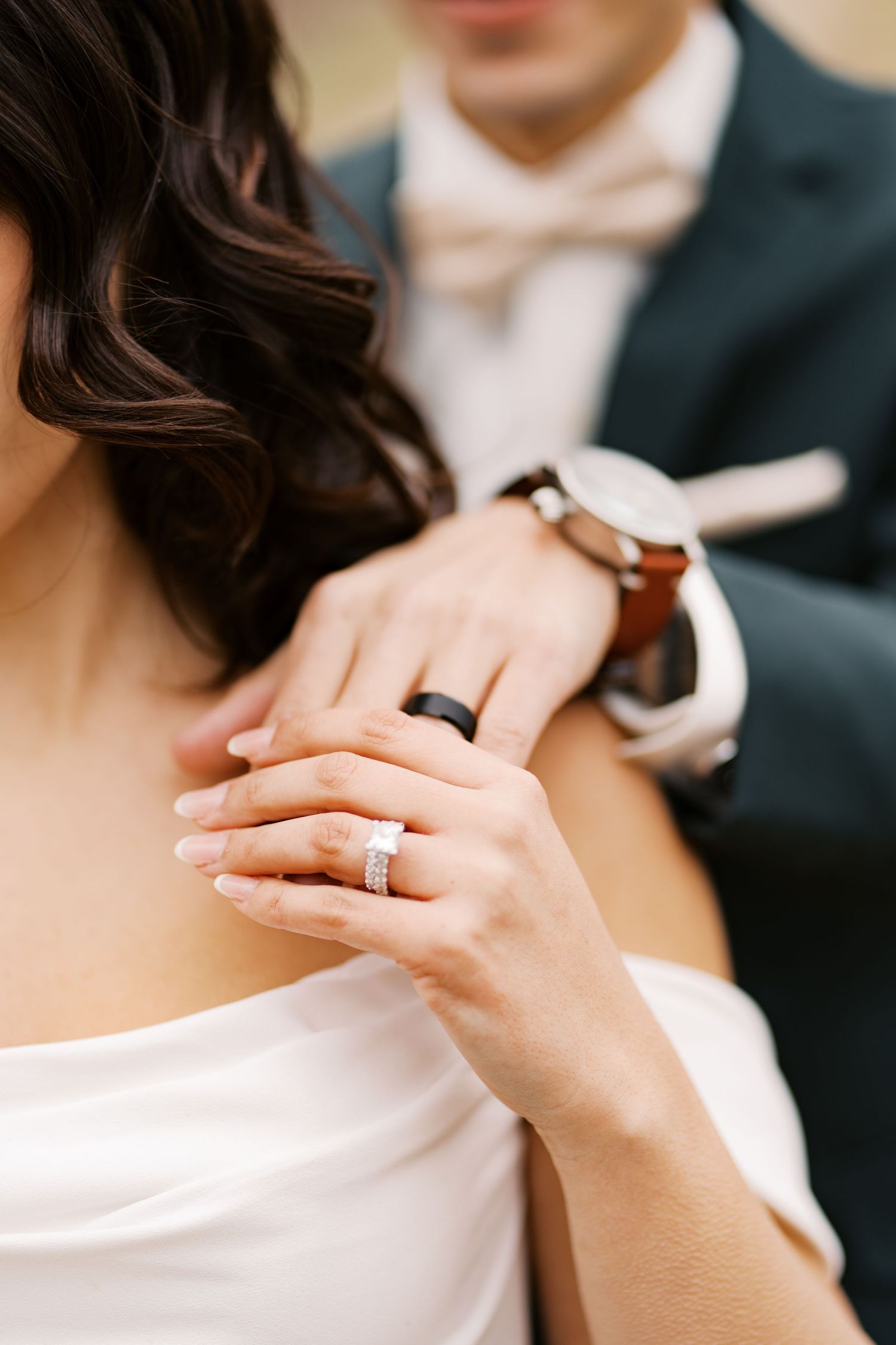 Detailed shot of a bride's hand resting on the groom's shoulder, showcasing a classic diamond engagement ring and black wedding band.