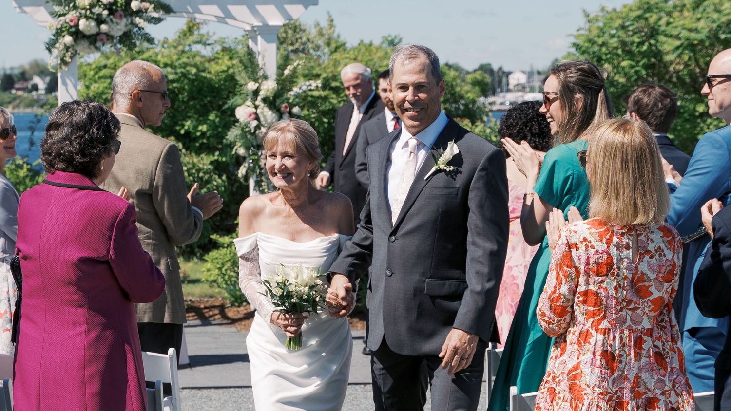 A bride and groom walking down the aisle together after their waterfront ceremony at The Boathouse in Tiverton, RI, while guests cheer.