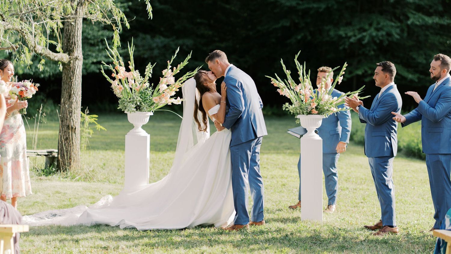 Outdoor wedding ceremony first kiss between a bride and groom at Salt River Farm, featuring white floral pillars and a forest backdrop.