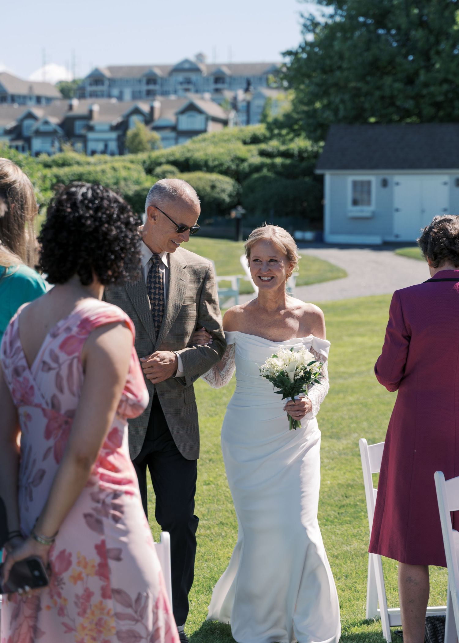 A bride in a classic off-the-shoulder gown walking down the outdoor aisle toward the Sakonnet River at her Tiverton, RI wedding.