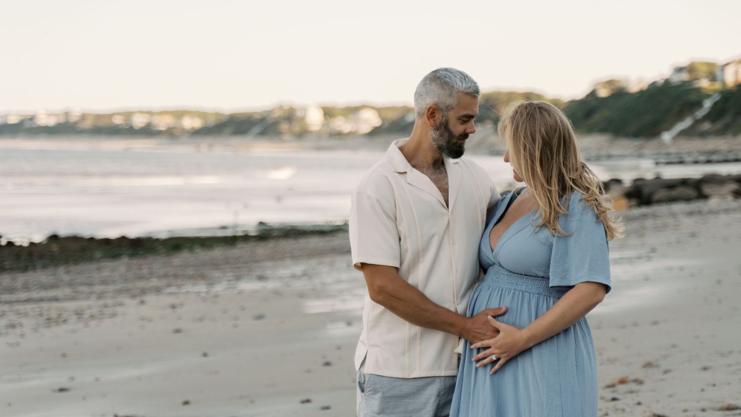 A posed photo of a man holding his wife's pregnant belly, taken under the bluffs at White Horse Beach in Plymouth, MA.
