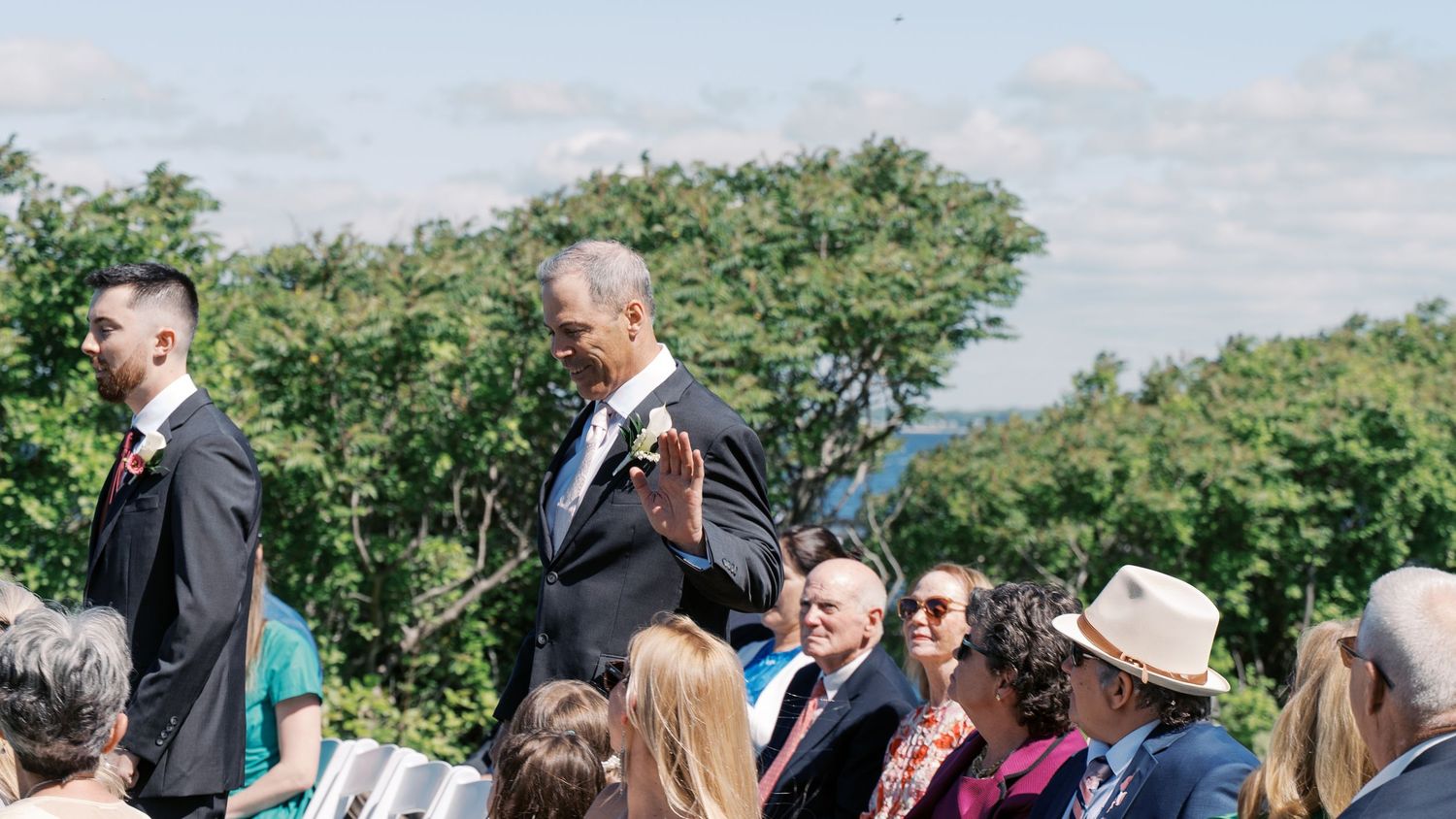 Candid shot of the groom waving to guests while walking down the aisle at a waterfront New England wedding celebration.