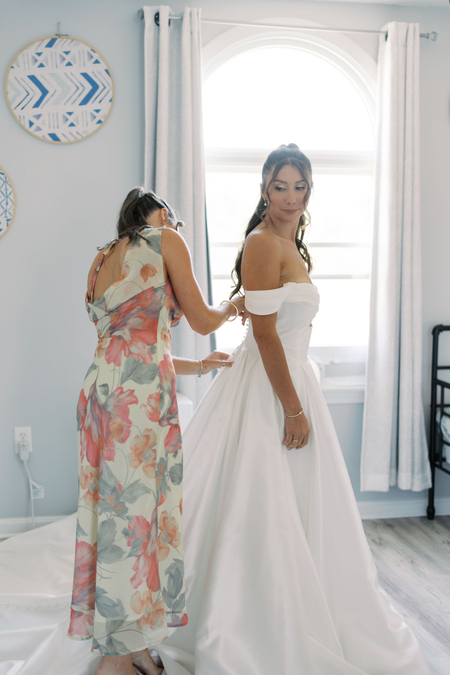 Candid moment of a bride in an off-the-shoulder gown being helped by her bridesmaid at a South Shore barn wedding.