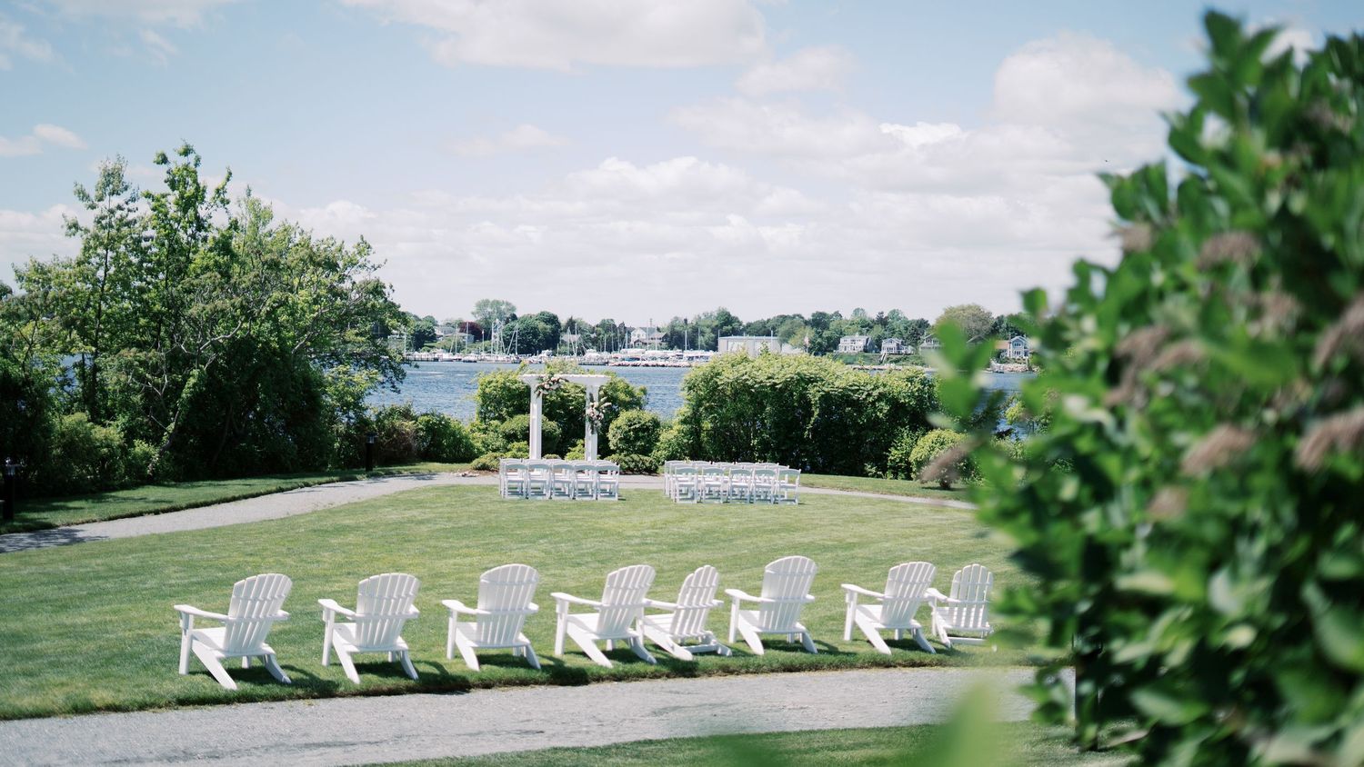 Outdoor waterfront wedding ceremony setup at The Boathouse featuring white Adirondack chairs and a garden arbor overlooking the bay in Tiver