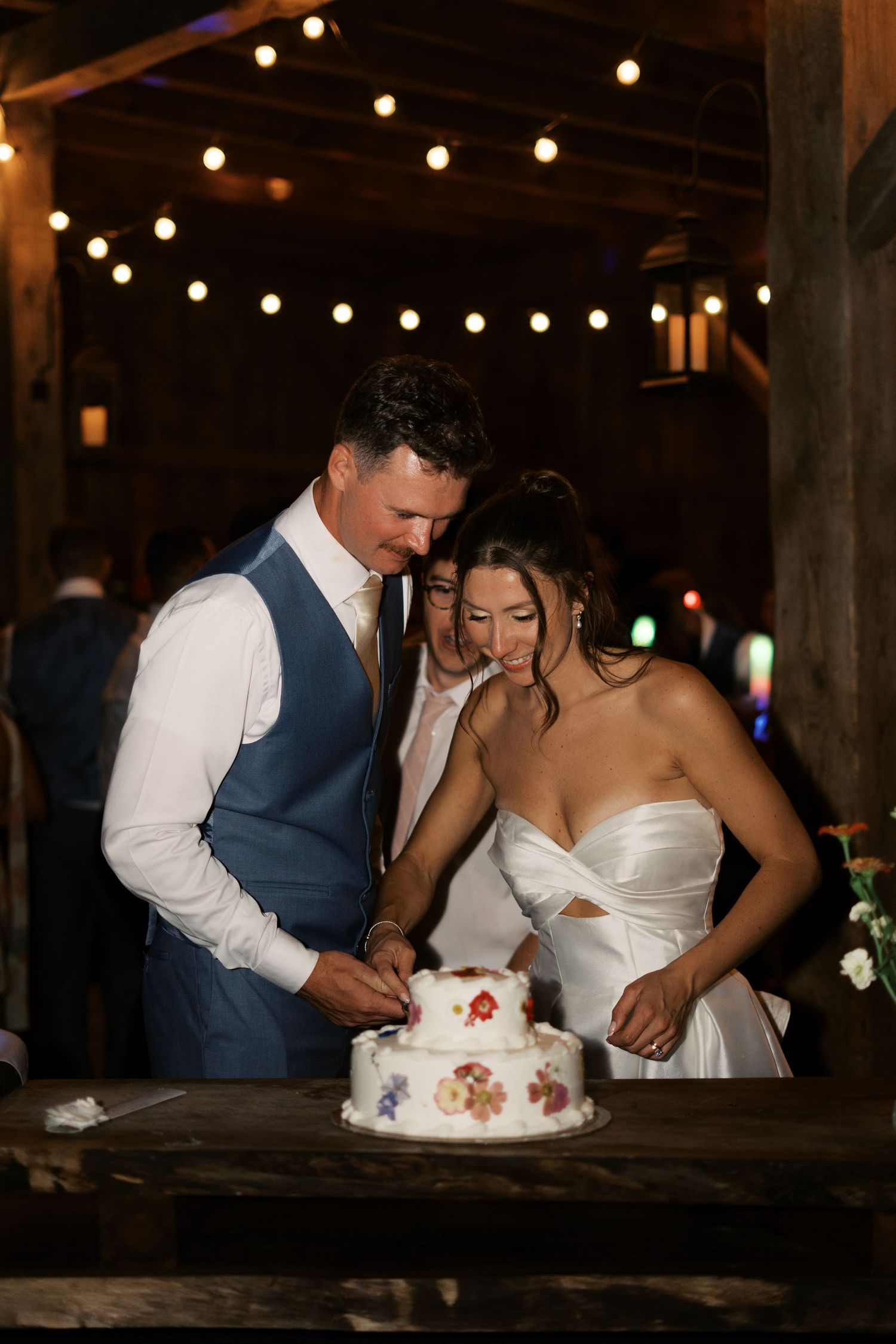 Newlyweds cutting a minimalist two-tier wedding cake decorated with pressed wildflowers at a rustic New England barn reception.