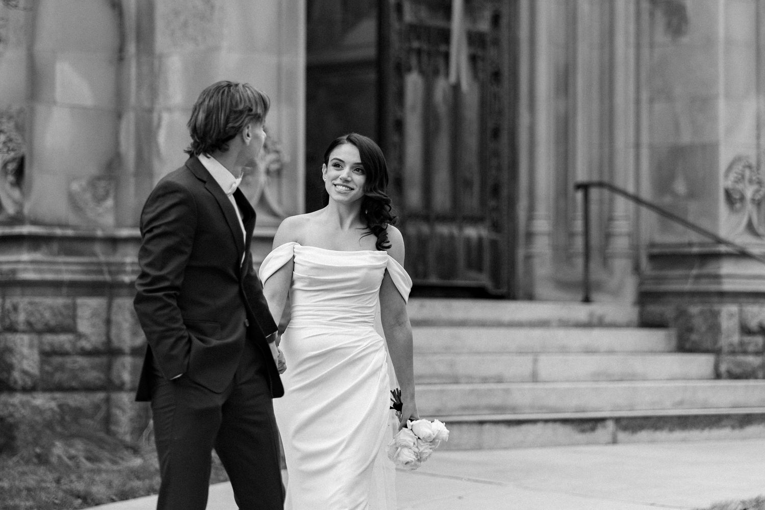 Black and white wedding portrait of a bride & groom walking outside the Gothic stone architecture of Unitarian Memorial Church in Fairhaven.