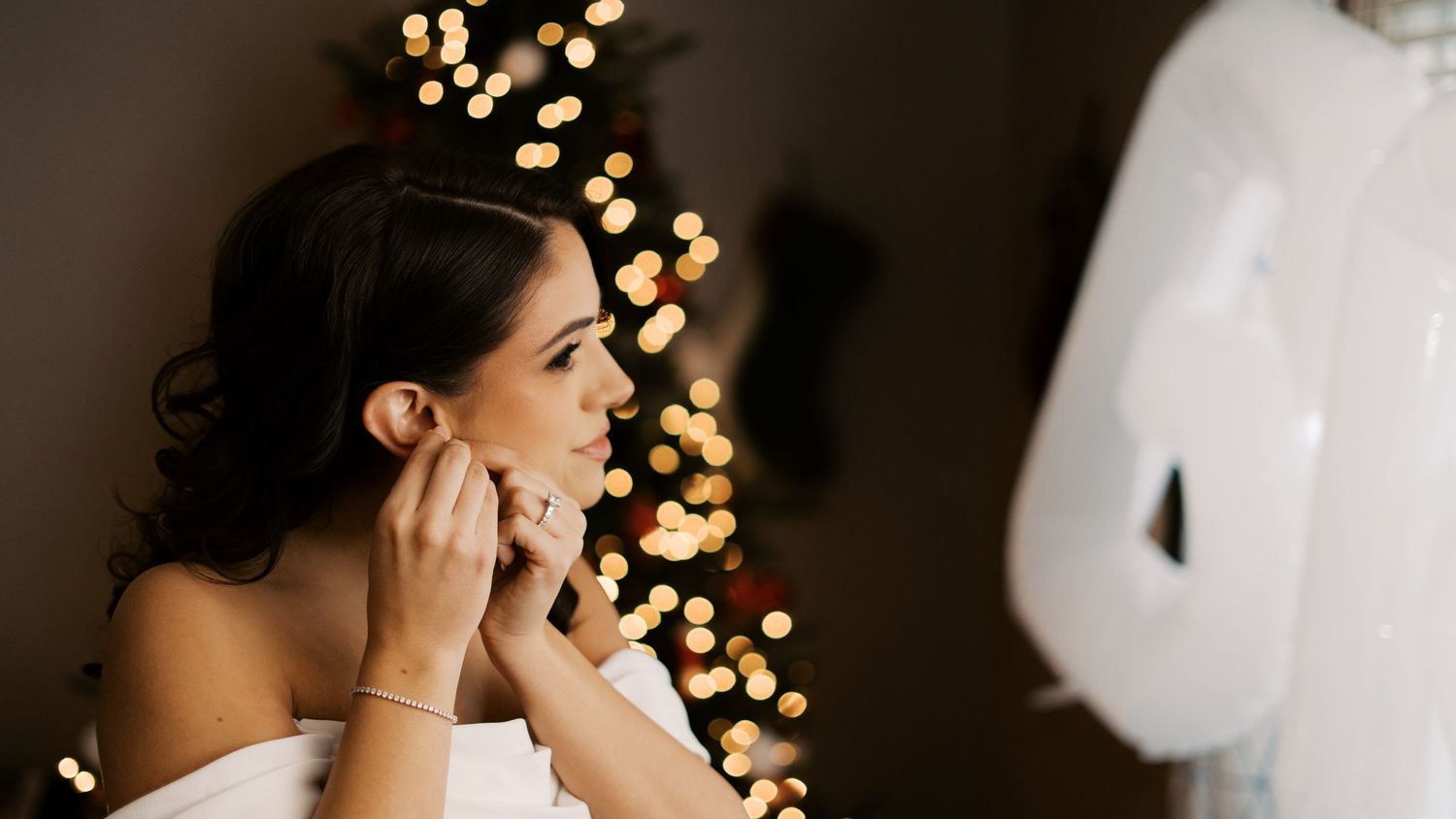 A bride putting on her earrings in front of a warm, blurred Christmas tree light background during her winter wedding prep.