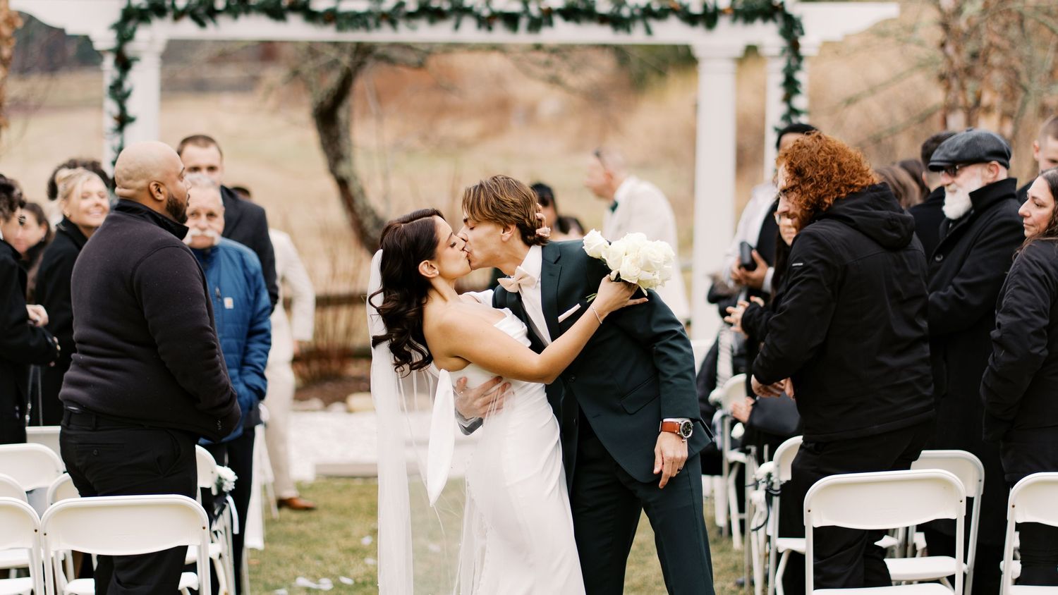 Bride and groom sharing a dramatic first kiss while guests applaud during a winter outdoor ceremony at Rosebrook Event Center.