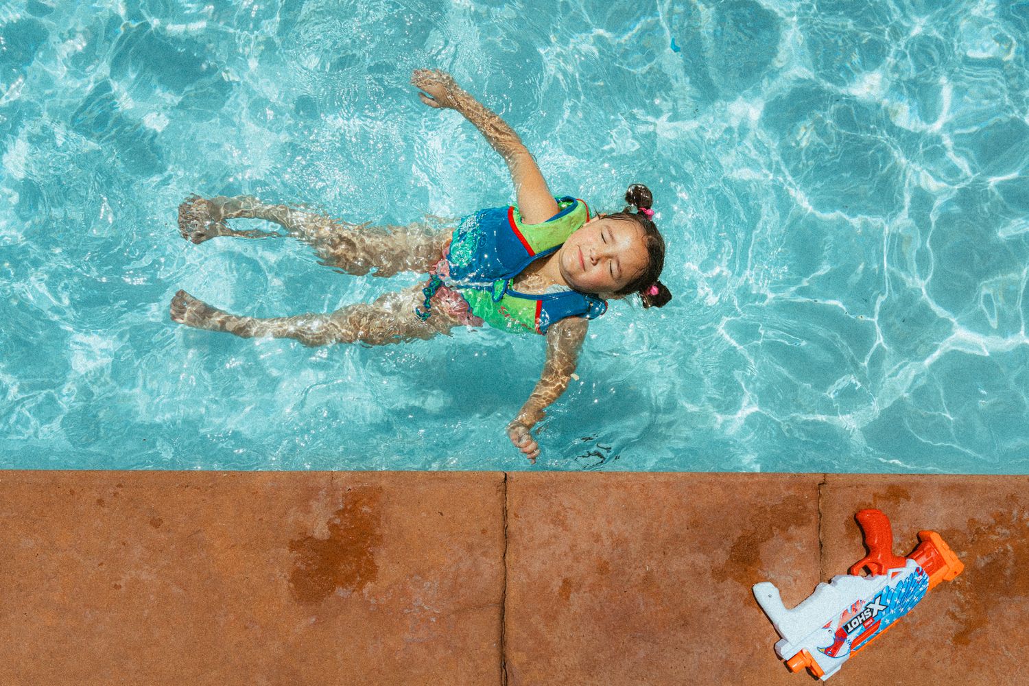 Young girl swimming in a clear blue pool wearing a colorful swimsuit, with a water gun toy resting on the poolside.