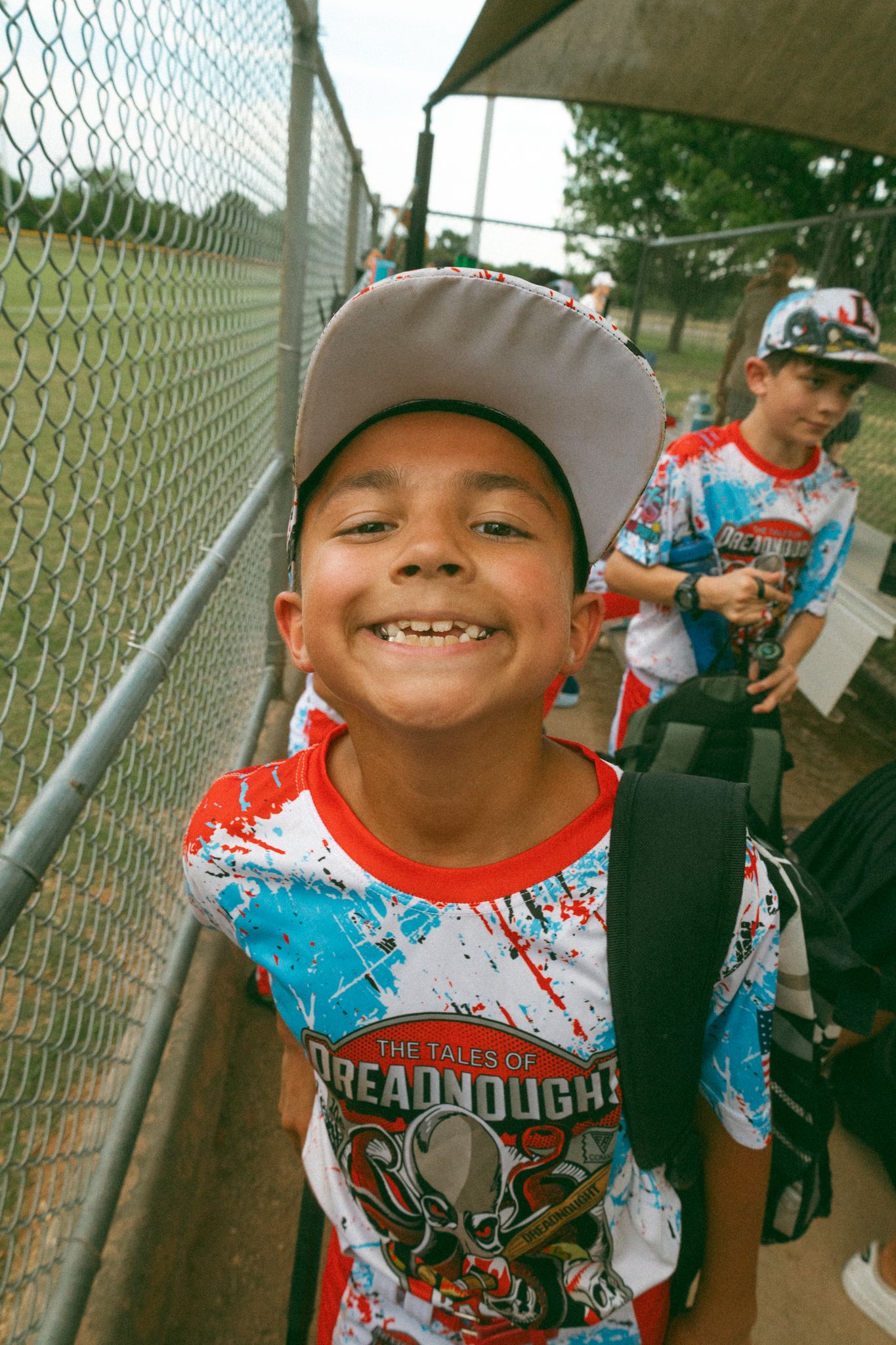 Smiling young boy in a baseball cap and Dreadnought shirt stands near a chain-link fence at a youth baseball game.