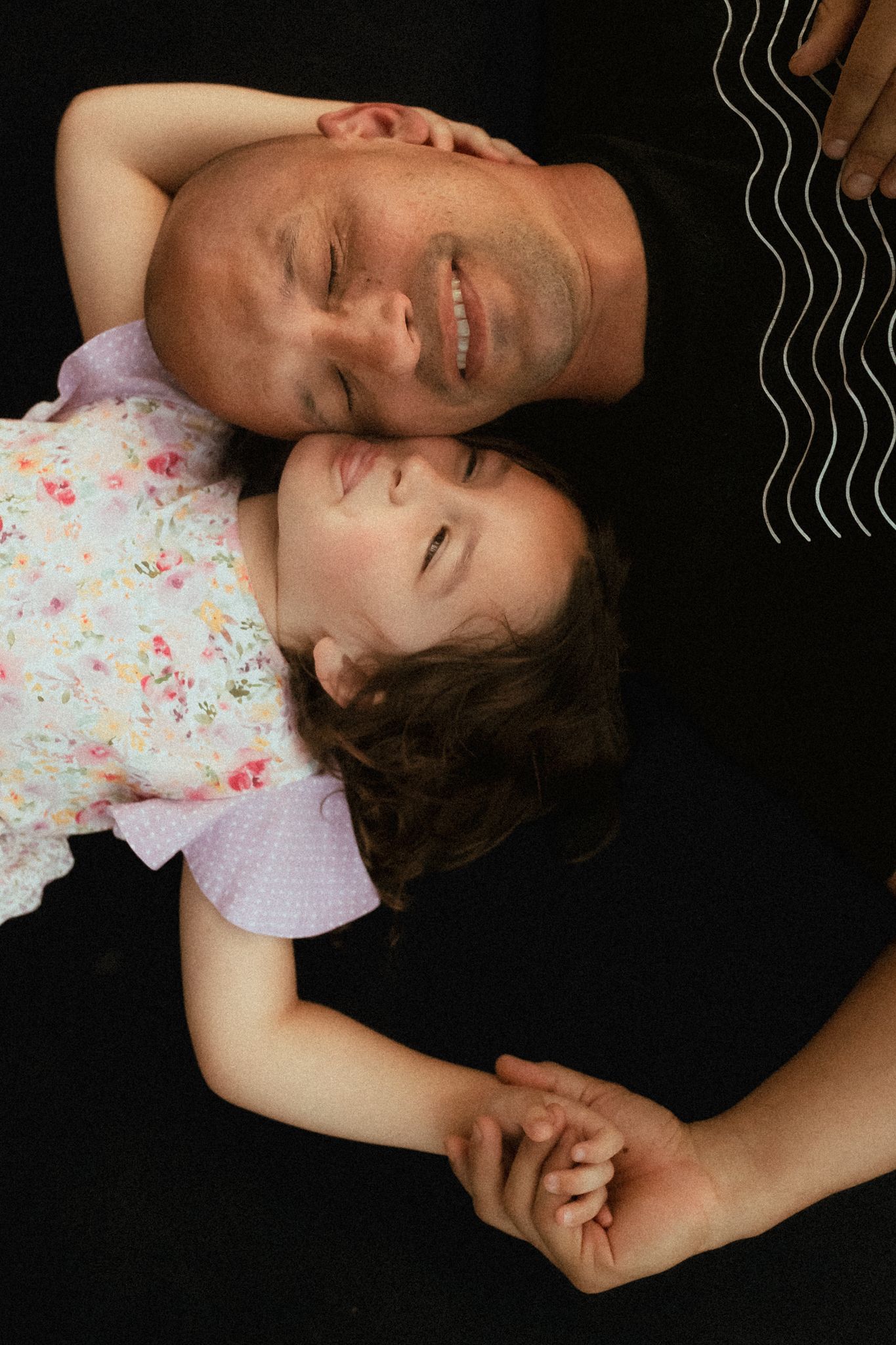 A smiling father and daughter lie together on a dark floor, holding hands in a tender moment.