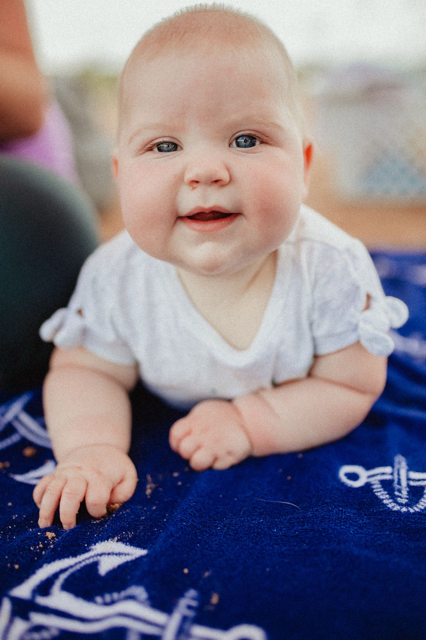 Baby in white outfit smiling while lying on blue patterned blanket.