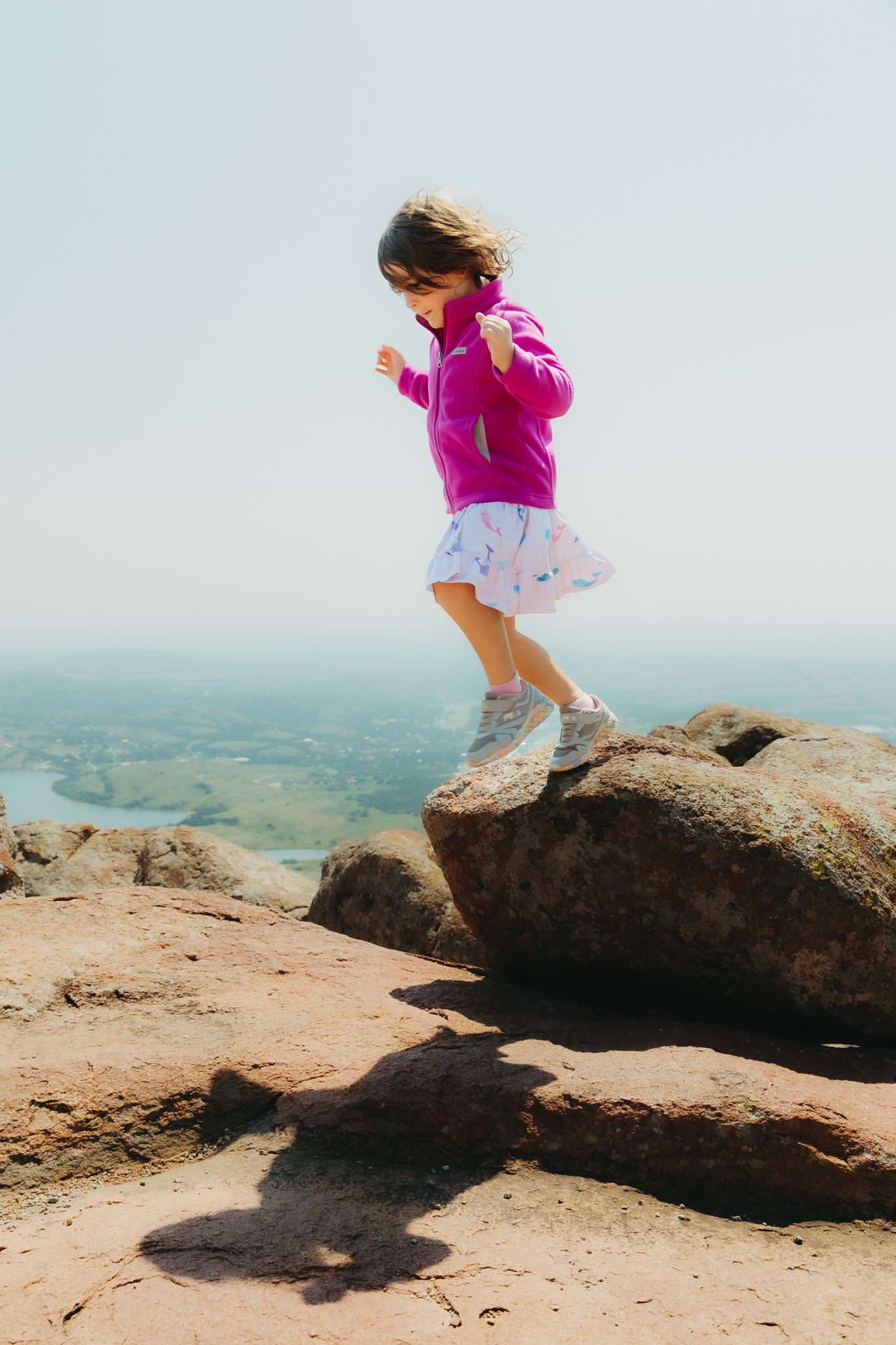 A young figure in a bright pink jacket leaps across rocky terrain on a mountaintop overlook with scenic views below.