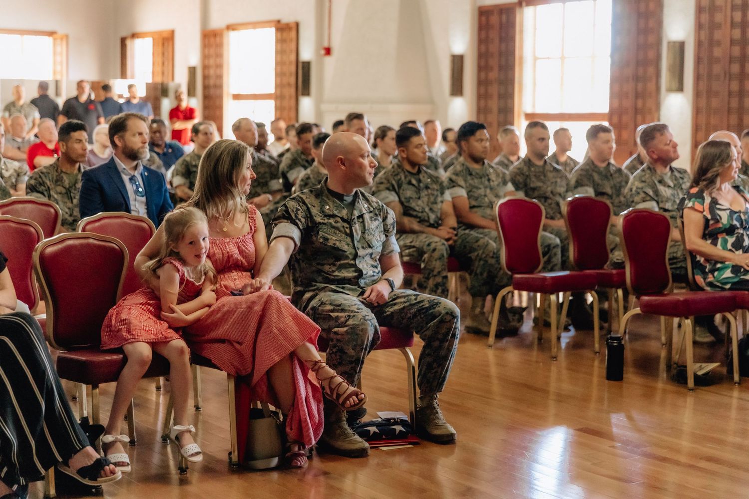 Military personnel and families seated in a formal hall during a ceremony, with soldiers in camouflage uniforms filling rows of red chairs.