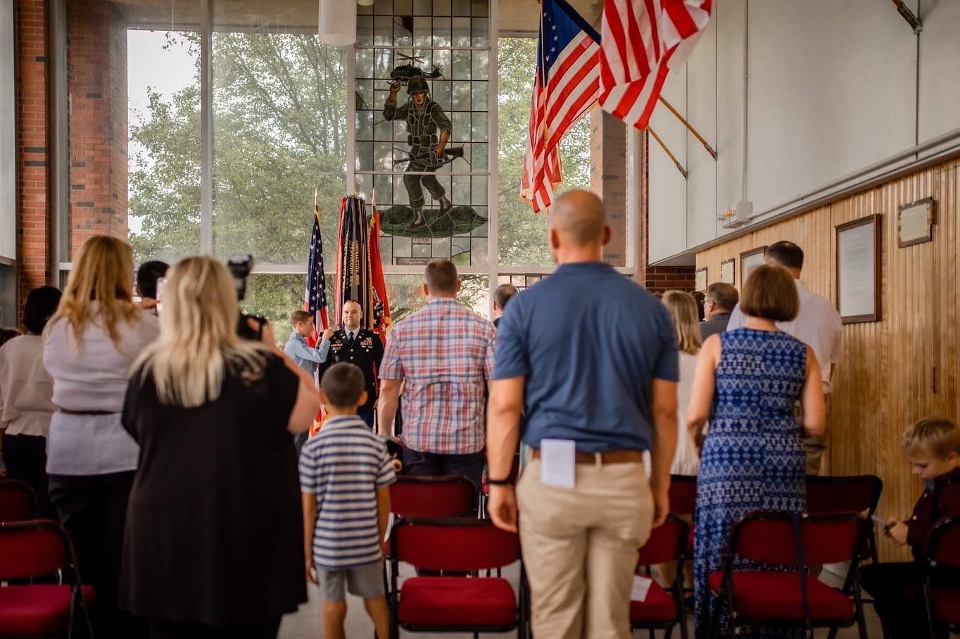 A crowd of people stand during a patriotic ceremony with American flags displayed in a wooden interior space.