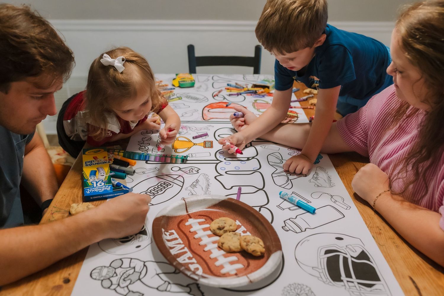 Family of four coloring together at a wooden table with Crayola markers and cookies on a plate.