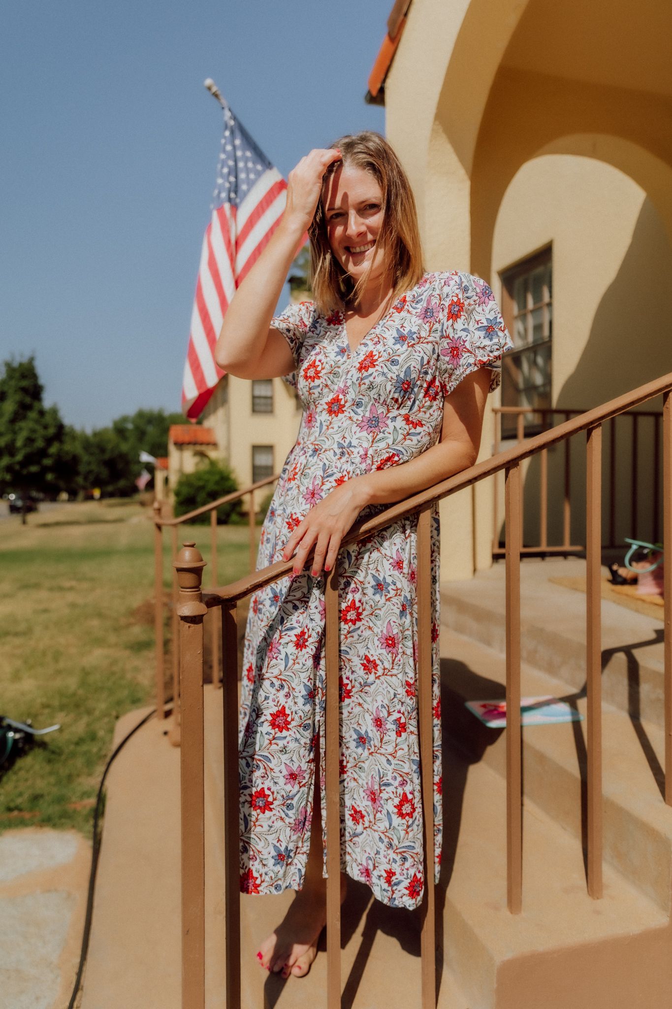 A woman in a floral print dress stands on a porch with American flags in a series of casual summer poses.