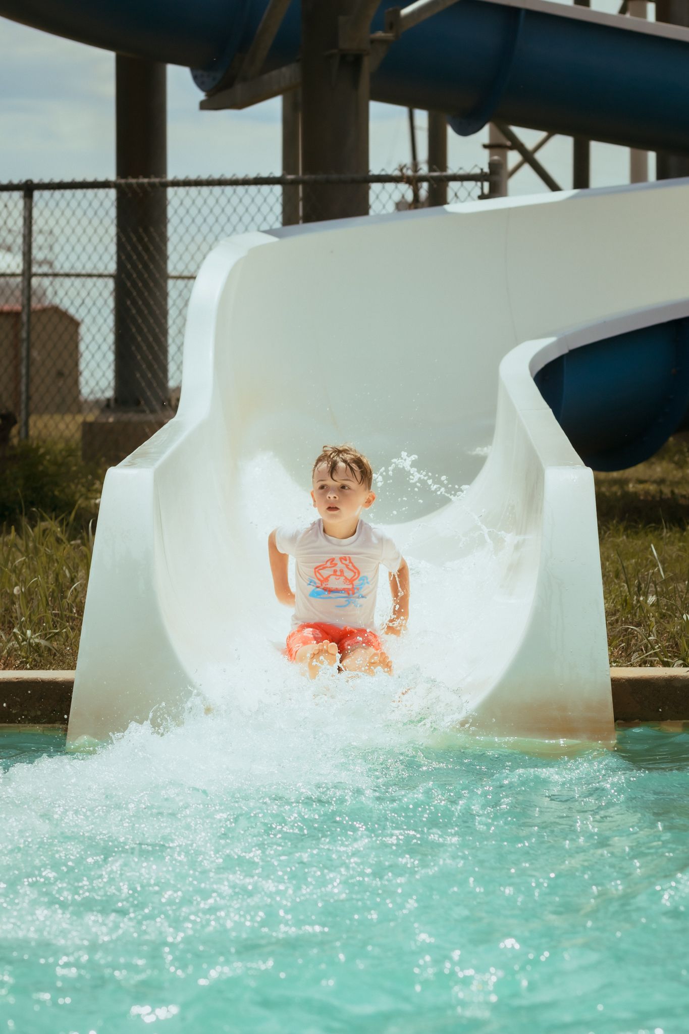 Child sliding down white water slide with joyful expression at outdoor pool.