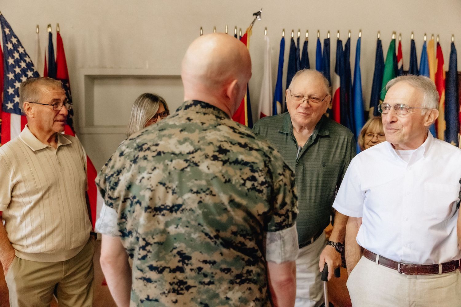Military officer in camouflage uniform speaks with civilians near international flags at a formal indoor gathering.