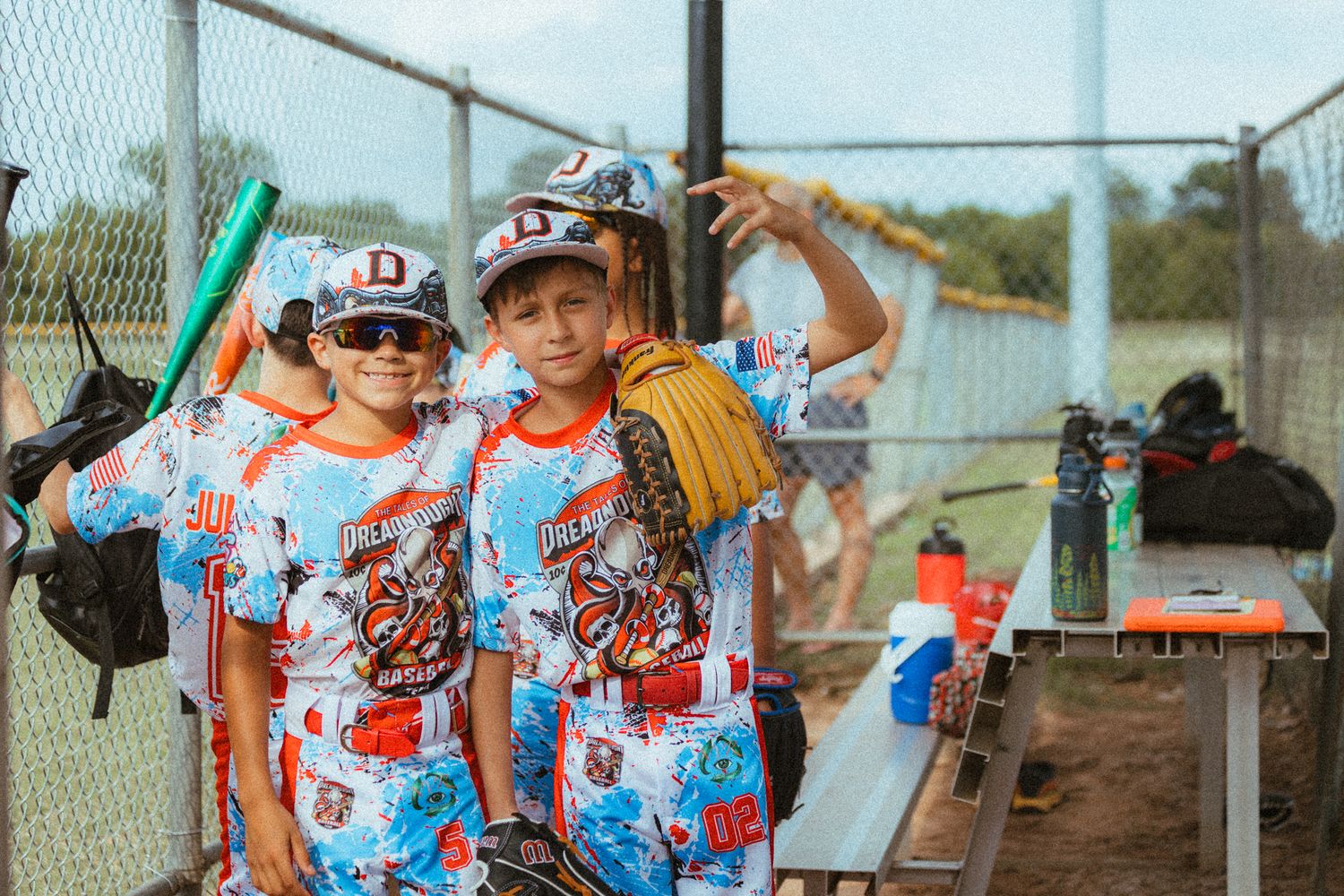 Two young baseball players in colorful uniforms pose together in the dugout holding gloves and bats.