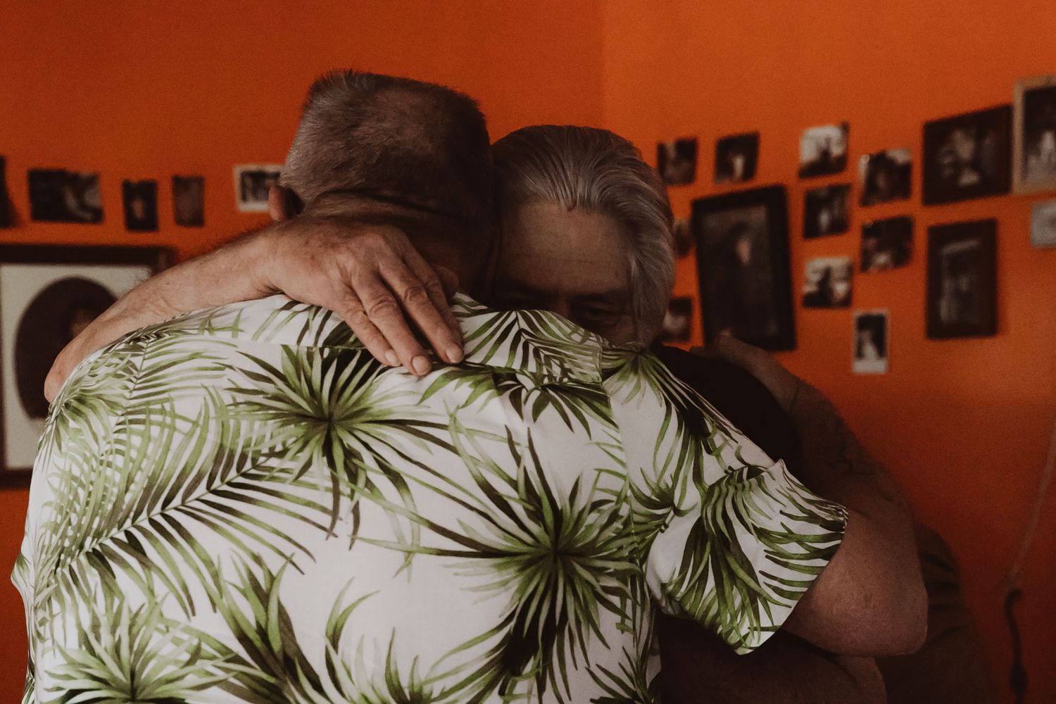 Two people embracing while wearing casual attire against orange wall with photos.