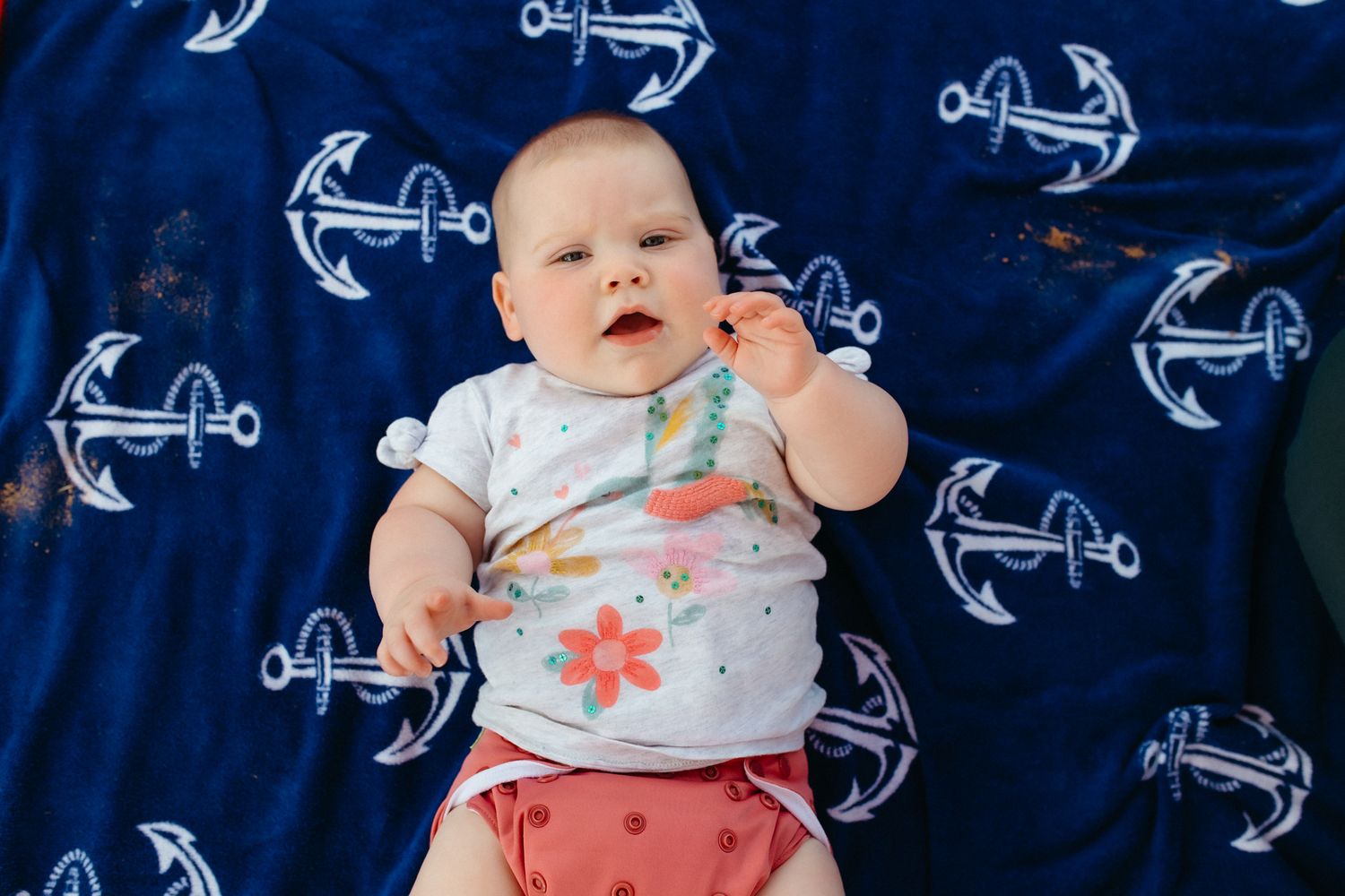 Baby lying on blue anchor-patterned blanket wearing floral outfit.