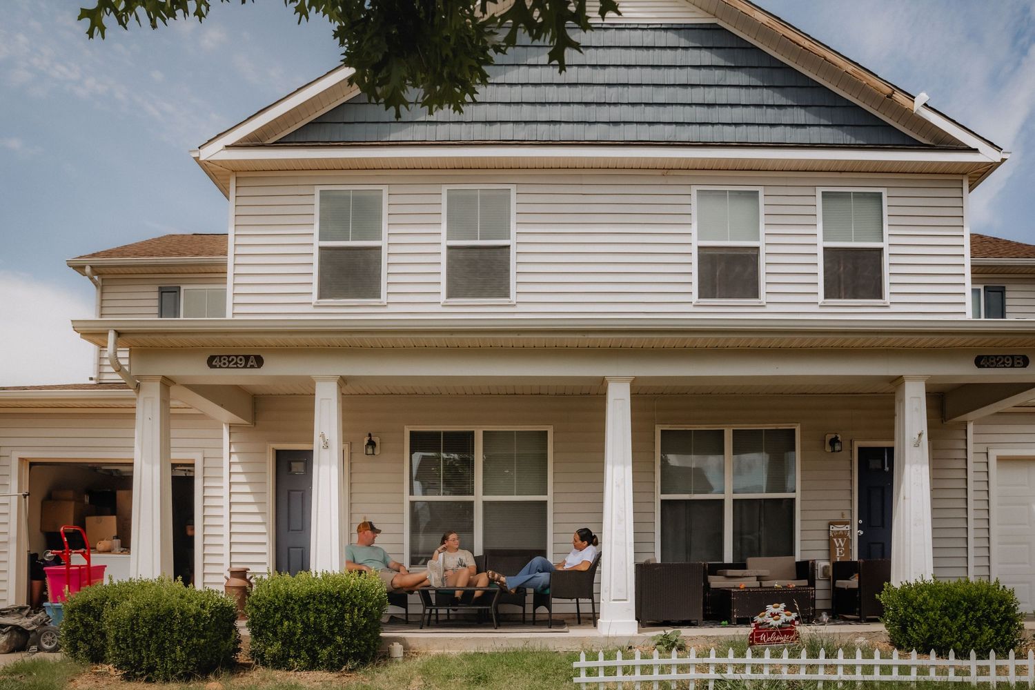 People relaxing on the front porch of a charming two-story beige home with white trim and a dark roof.