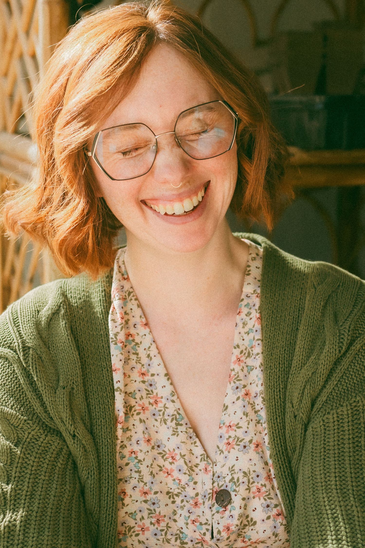 A joyful portrait in warm lighting showing someone wearing round glasses, a green cardigan and floral print blouse.