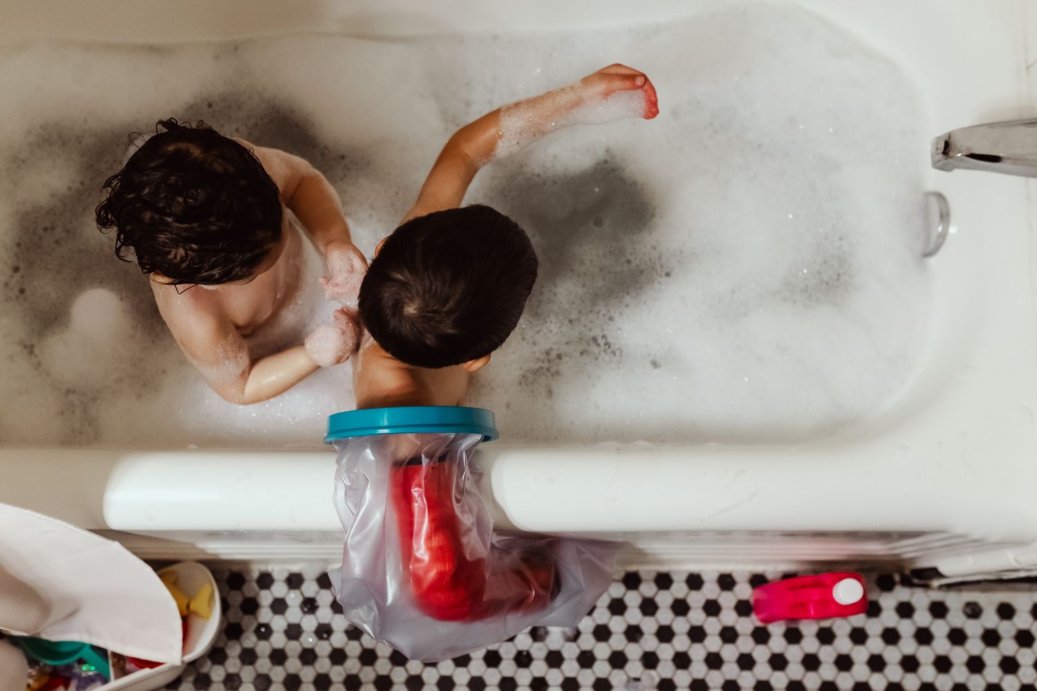 Two young children playing with toys in a bubble bath, viewed from above in a white bathtub.