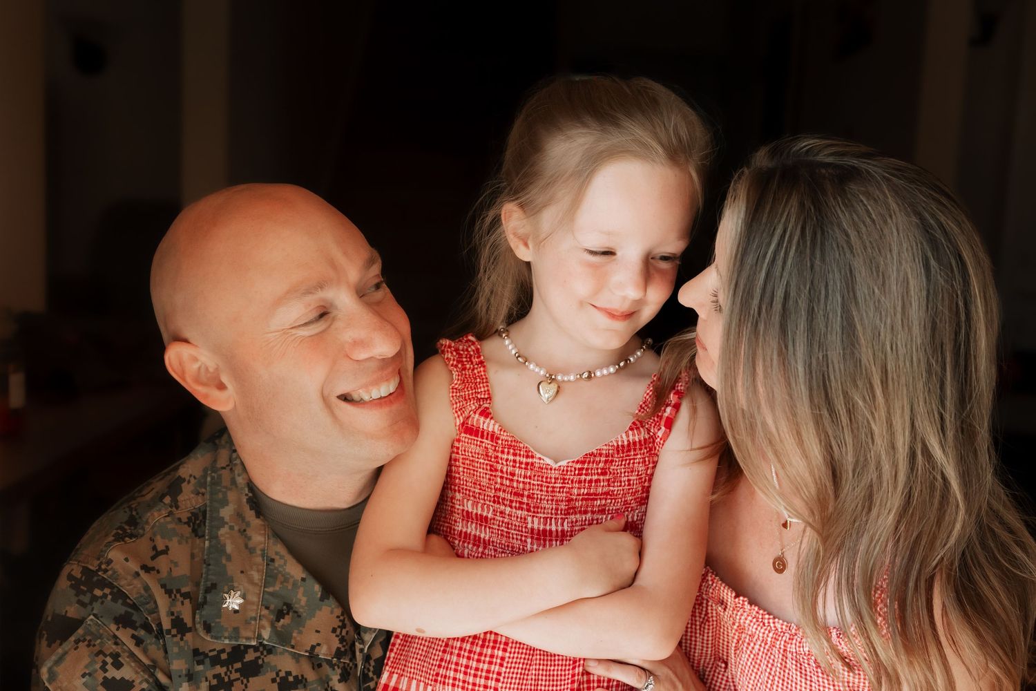 A smiling bald man in military uniform holds a young girl in a red dress alongside a woman in a warm family portrait.