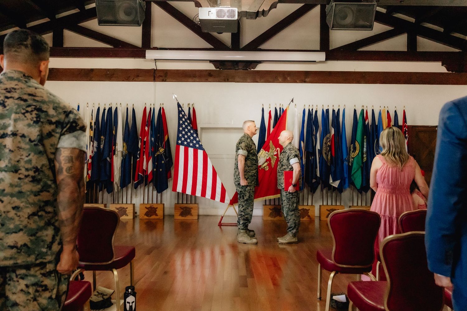 Military ceremony in a hall with US and colorful flags, two Marines exchanging a guidon while others watch.