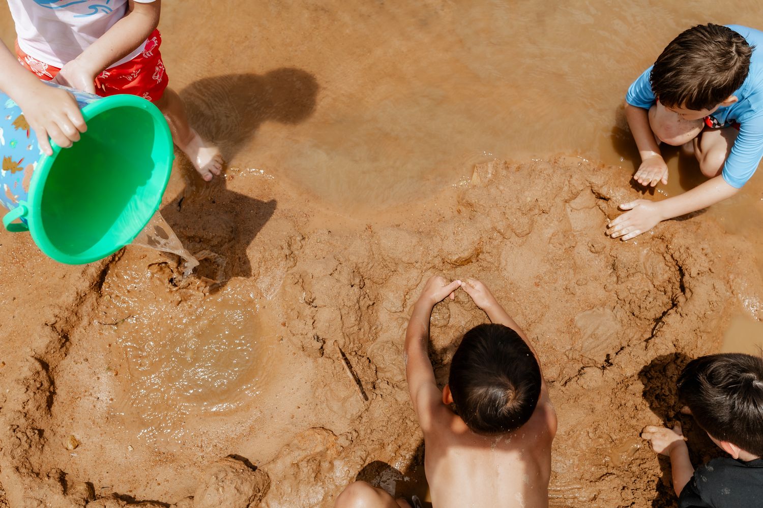 Children playing with buckets and toys in muddy sand near the water.