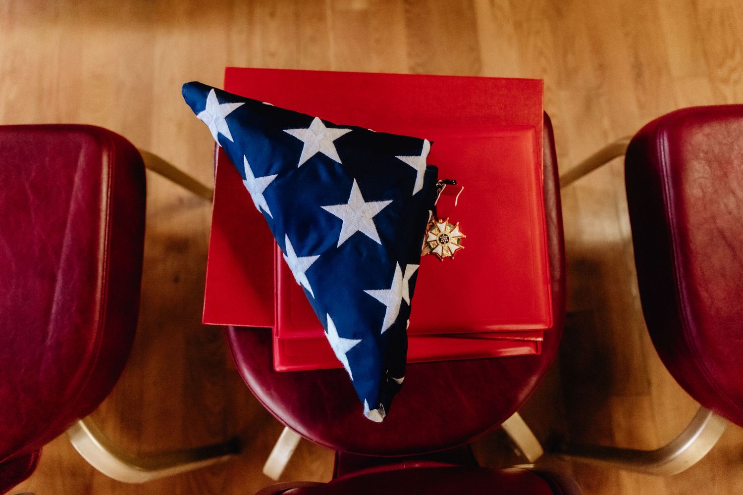 Folded American flag with gold pin resting on red book between two red chairs.