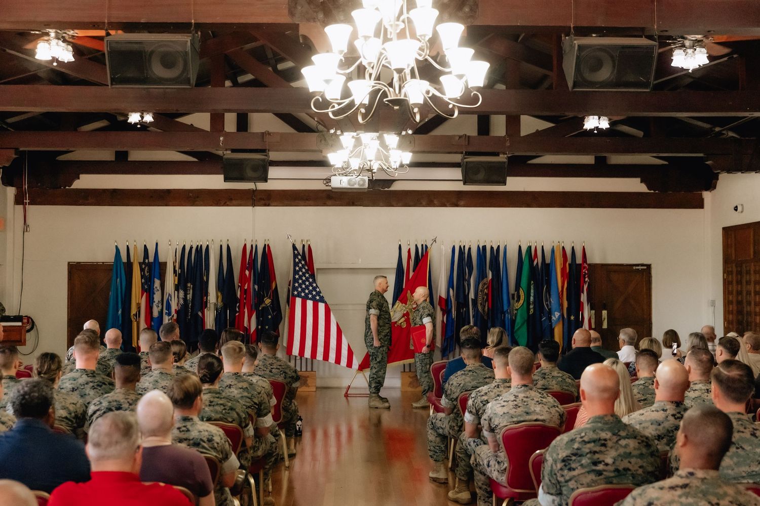 Military ceremony in a decorated hall with soldiers seated before a speaker, flanked by colorful flags and a chandelier above.