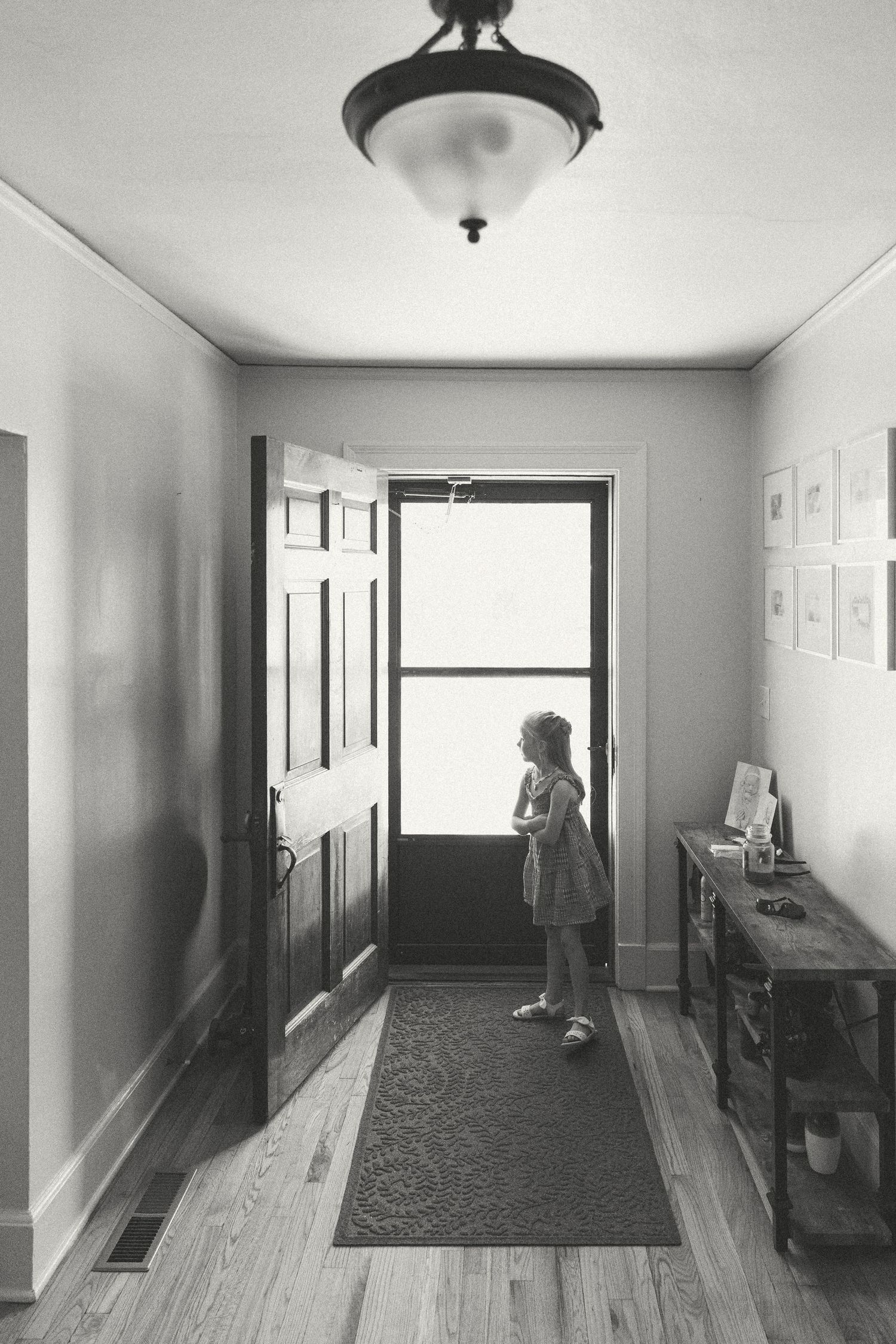 Black and white photo of a young girl standing in a hallway looking out an open front door.
