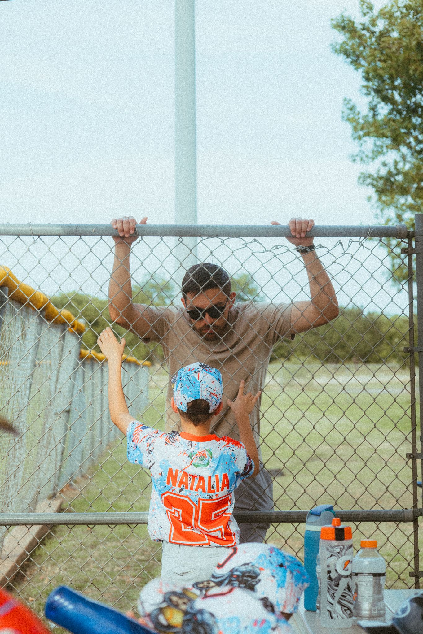 A man holds a chain-link fence while a child in a Natalia baseball jersey stands beside him at a sports field.
