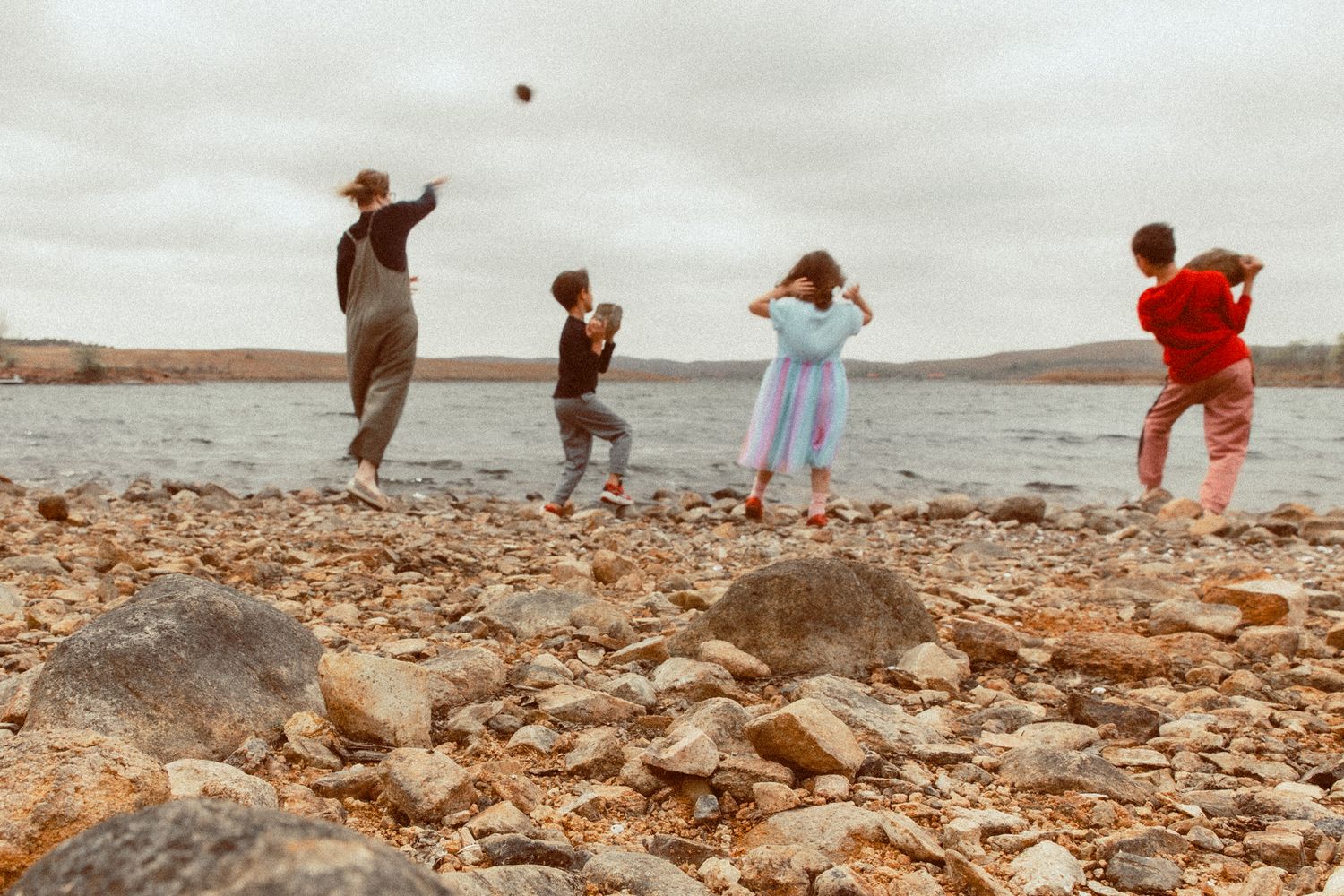 Four children skipping stones along a rocky lakeshore on an overcast day.
