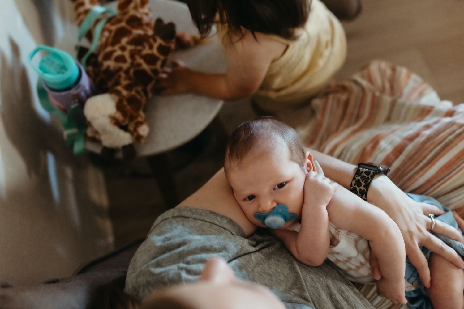 A baby in a diaper and pacifier lies on a couch cushion while someone sits nearby with a stuffed giraffe.