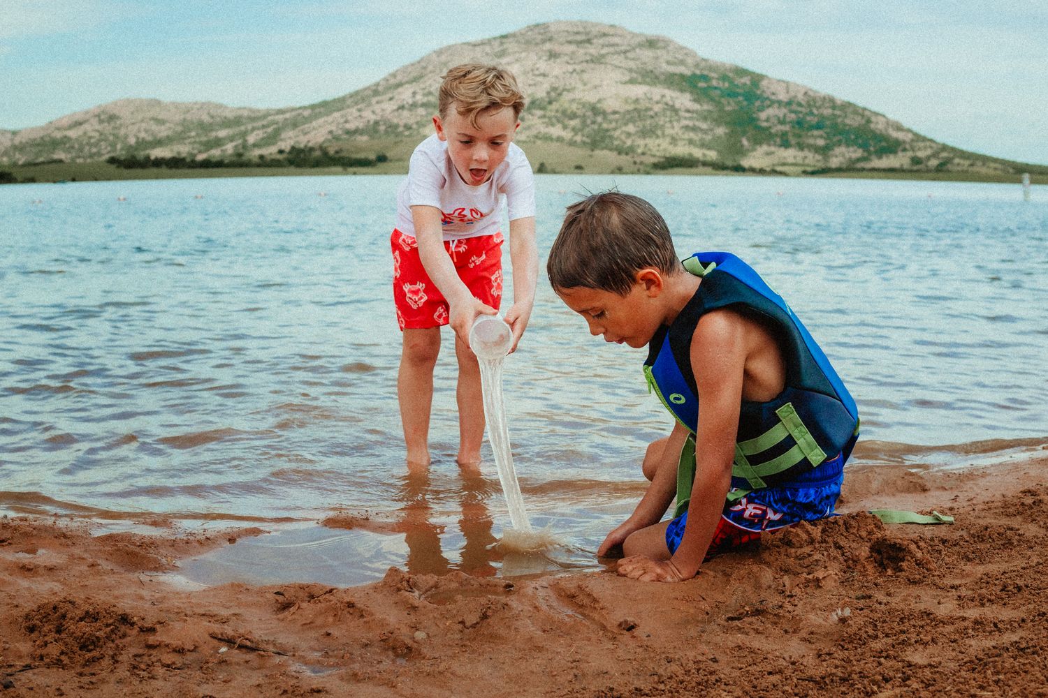 Two children playing in wet sand at the edge of a lake with mountains in background.