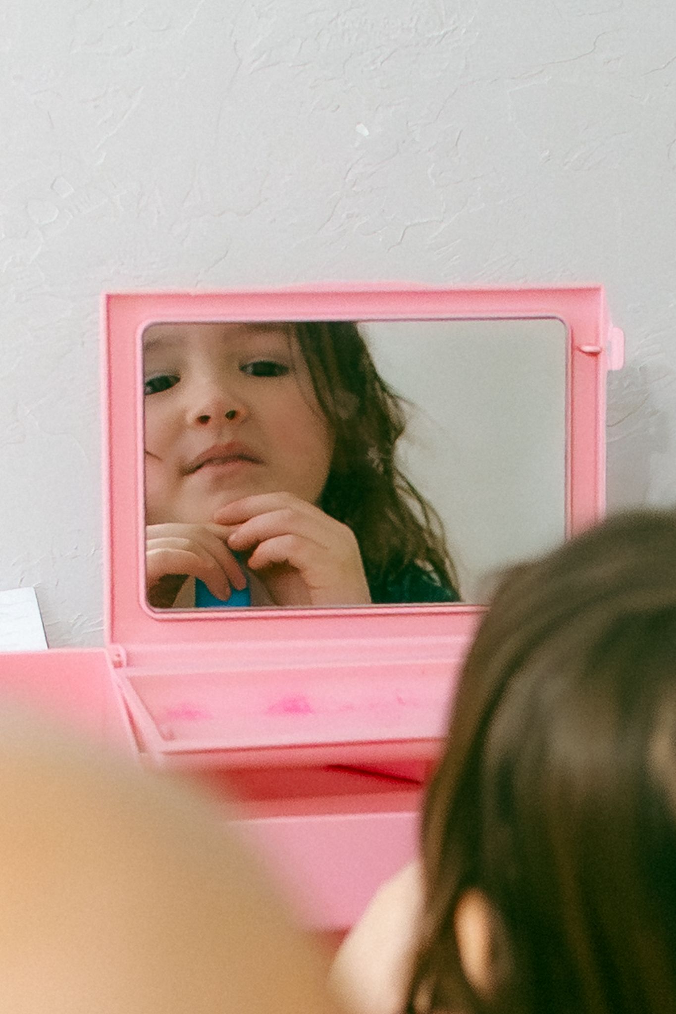 A pink-framed mirror reflects partial views of someone's face in a soft, dreamy aesthetic composition.
