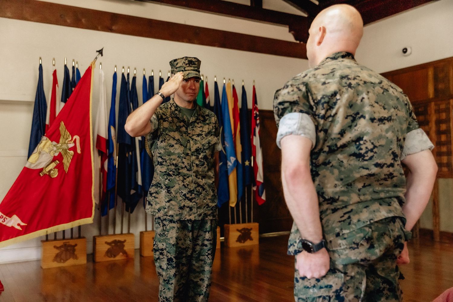Two U.S. Marines in camouflage uniforms exchange salutes indoors with military flags displayed in background.