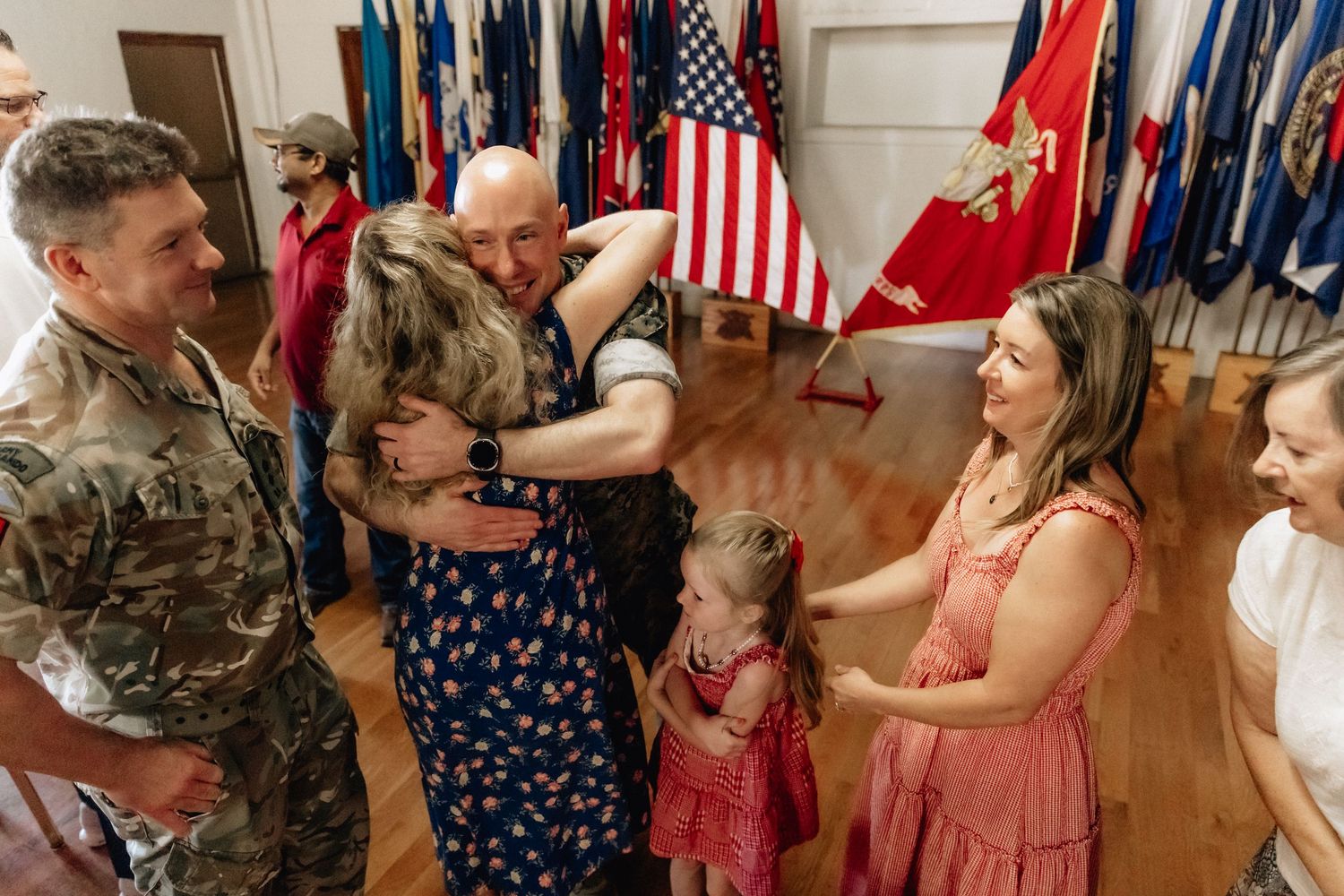 Military family reunion with soldier embracing loved ones amid American and Army flags in a ceremonial hall.