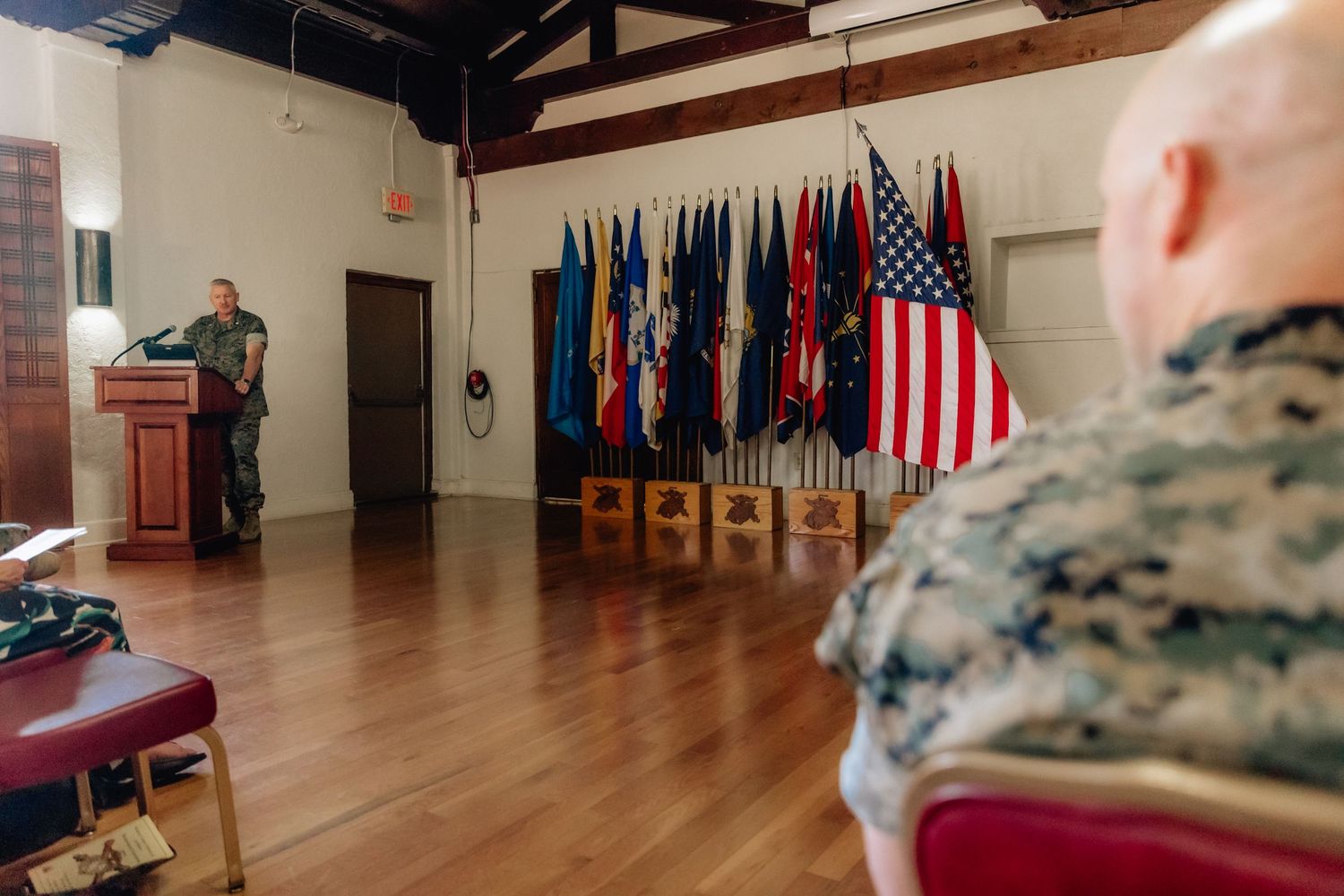 Marine officer speaks at podium before military flags during an official indoor ceremony with seated attendees.