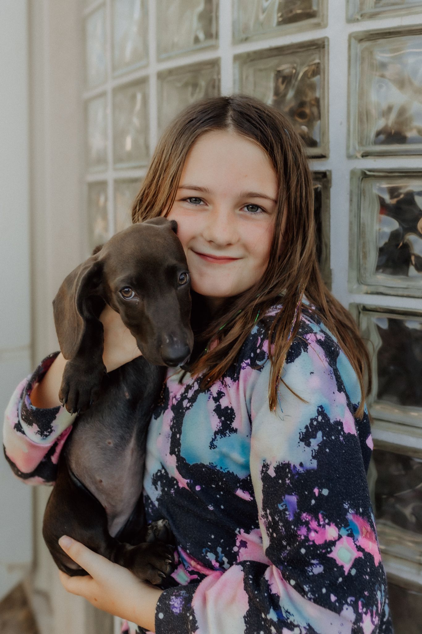 A smiling young girl in a colorful tie-dye shirt holds a black dachshund puppy against a glass block wall.